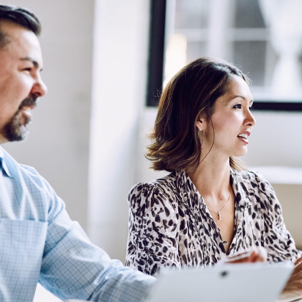 Two office workers sit side-by-side while looking at someone off-camera in a conference room setting
