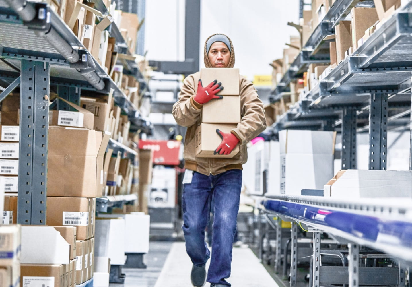 A warehouse worker wearing red gloves carrying three medium-sized boxes down an aisle. Boxes of various sizes line shelves on both sides of the worker.