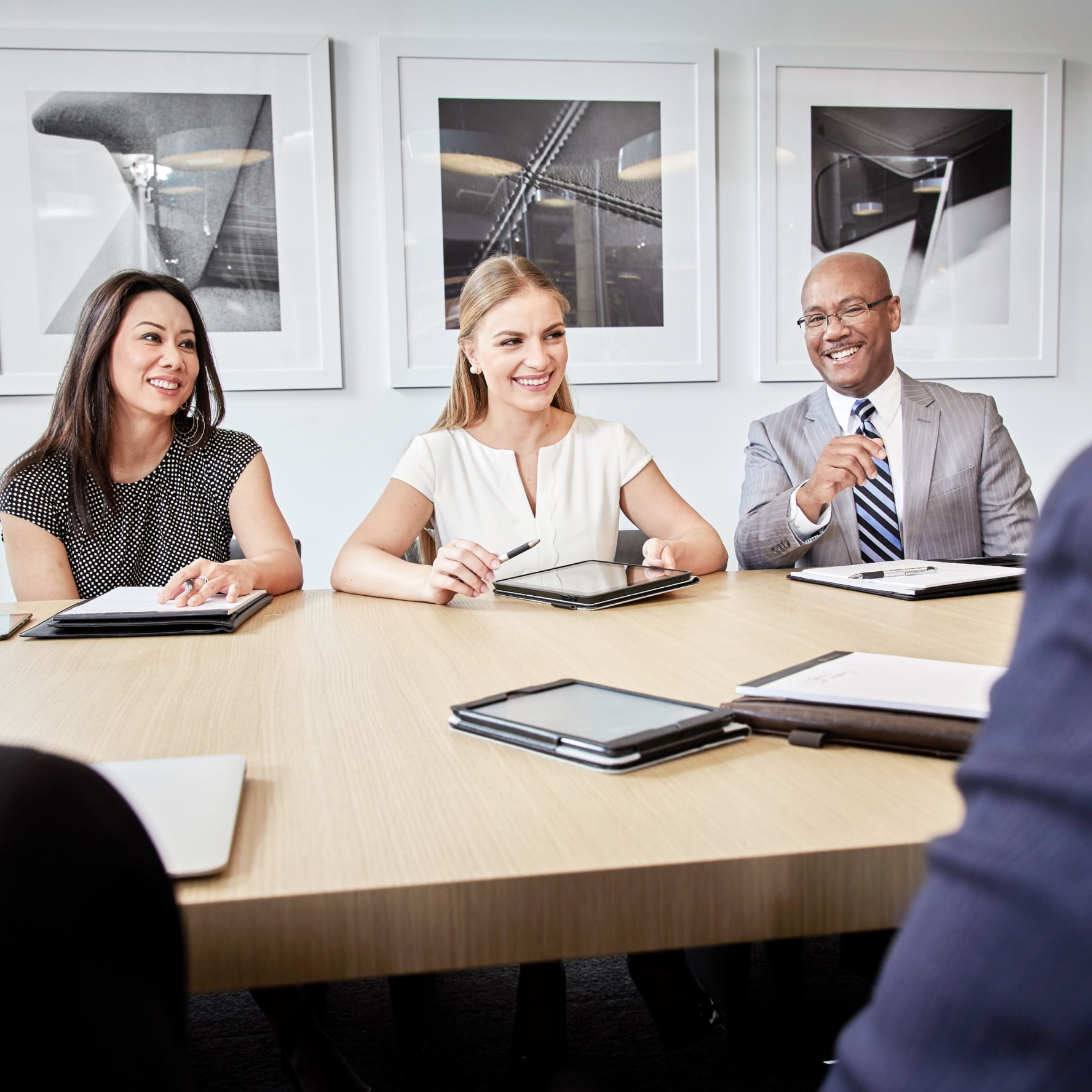 A diverse group of employees dressed in business clothing sitting at a table in a meeting room. Tablet devices, notepads, and pens sit on the table.