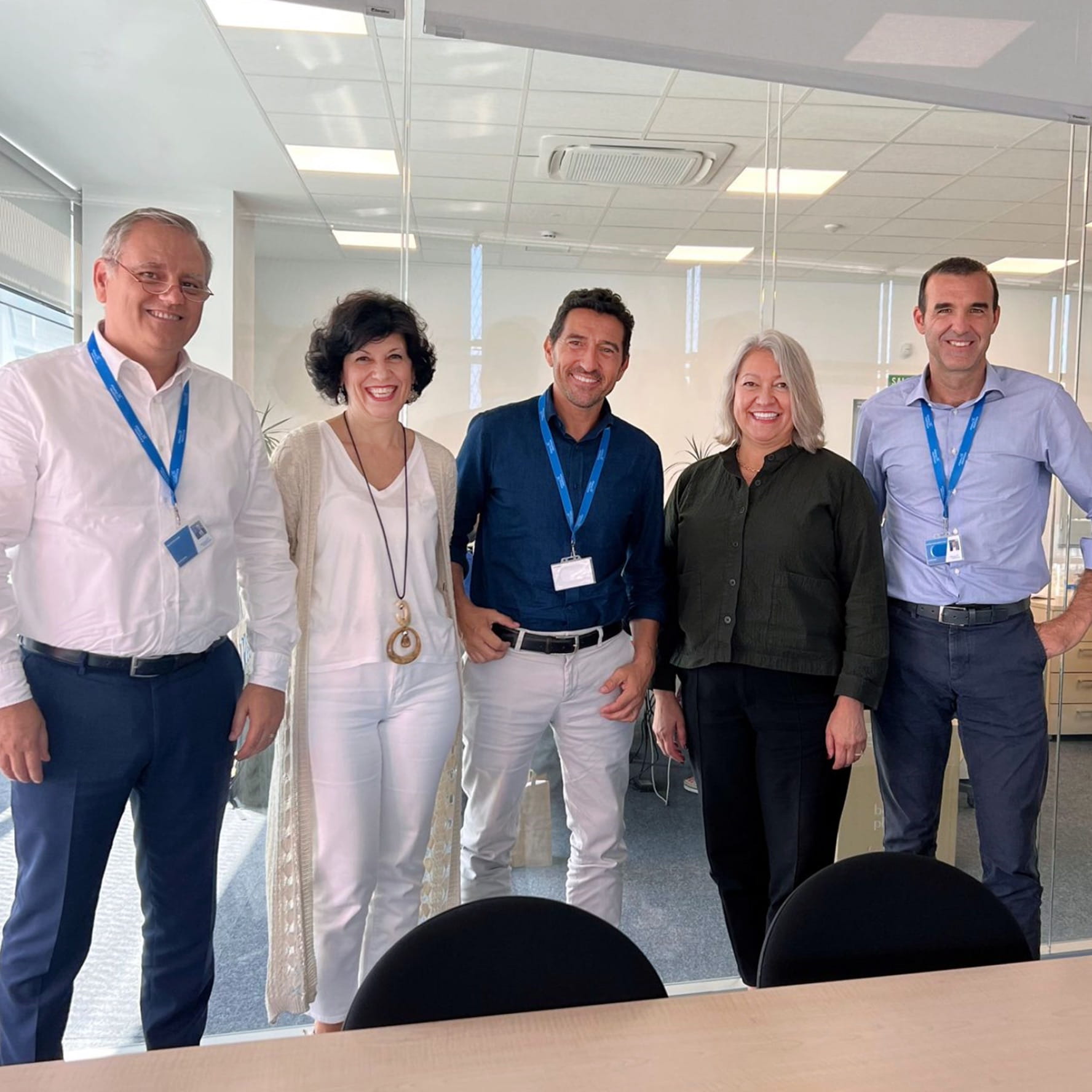 Five smiling Cencora employees standing side-by-side in an office conference room. They are dressed in business casual attire and three of them wear lanyards with name tags attached.