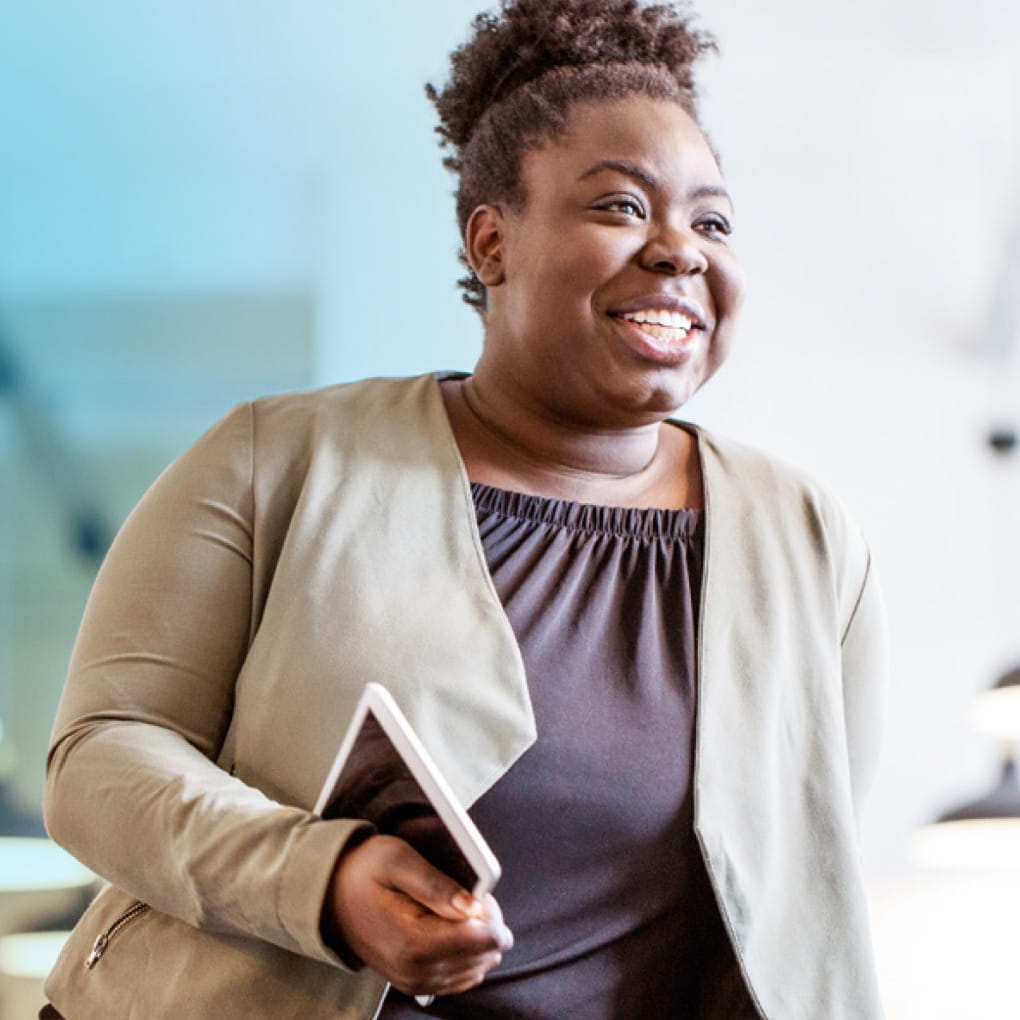 A dark-skinned person wearing business-casual clothing holding a tablet device while smiling.