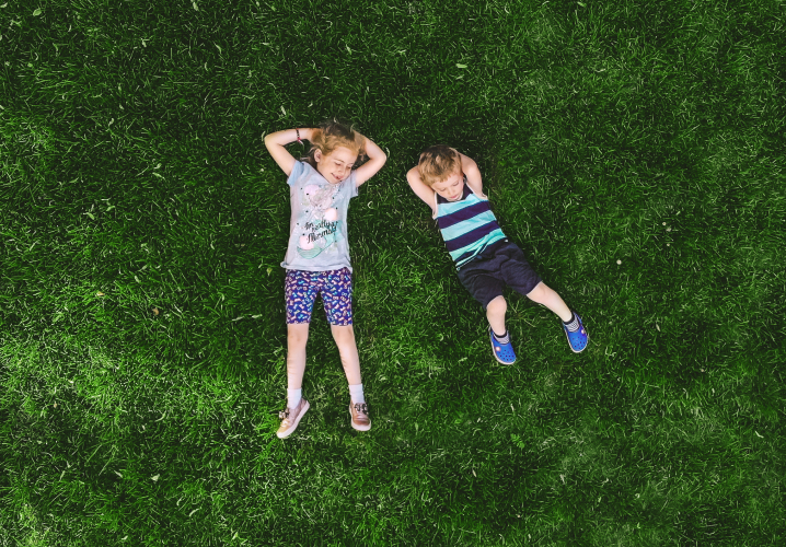 Two young children laying sprawled out side-by-side in a green lawn. They both have light skin, wear shorts and t-shirts, and have their hands behind their head.