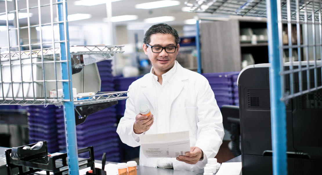 A light-skinned pharmacist holding a pill bottle while reading a sheet of paper at a pharmacy counter. They wear a white lab coat and glasses.