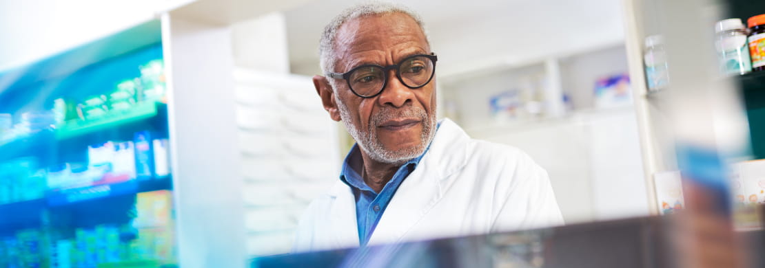 A dark-skinned pharmacist glances down at something out-of-frame as they work in a pharmacy. They wear a white lab coat and black glasses.