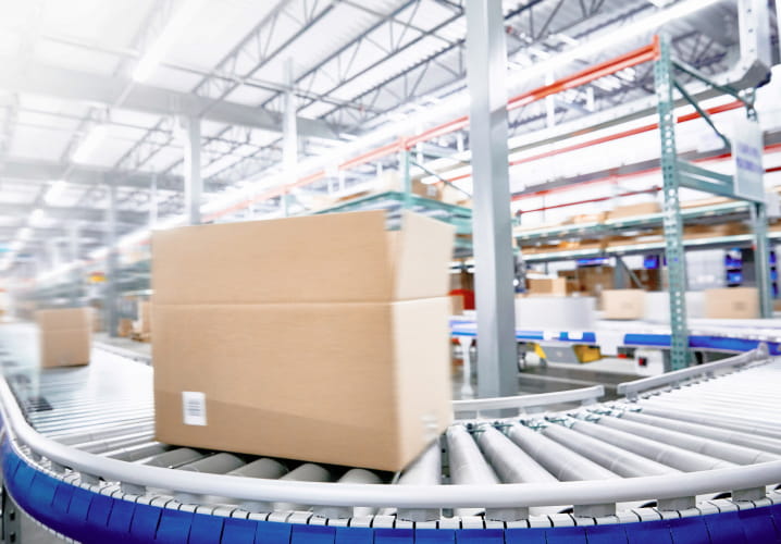 A large brown box moving down a conveyor belt inside of a large, bright warehouse factory.